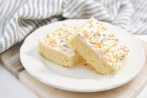 A white plate holds two Keto Sugar Cookie Bars topped with white frosting and colorful sprinkles. The plate is set on a white and wooden surface, with a striped cloth in the background.