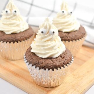 Three chocolate keto cupcakes with white frosting featuring edible googly eyes, resembling ghosts, sit on a wooden board. The cupcakes are in silver foil liners and the background includes a white cloth with a black grid pattern.
