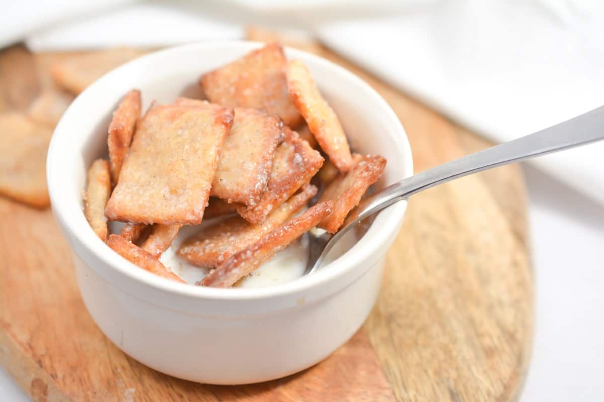 A white bowl filled with keto cinnamon toast crunch sits on a wooden board. The golden-brown crackers have a slightly rough texture and are sprinkled with coarse salt. A metal spoon is placed inside the bowl next to the crackers.