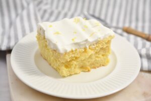 A slice of Keto Lemon Poke Cake topped with a generous layer of rich whipped cream and garnished with lemon zest on a white plate.
