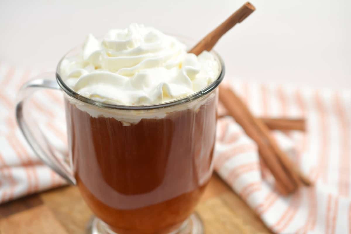 A close-up shot of a glass filled with Keto Apple Cider, topped with a generous swirl of whipped cream. A cinnamon stick is used as a garnish, leaning against the inside of the glass. The background features a soft, blurred fabric with light pink and white stripes.