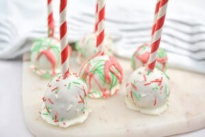 Five keto cake pops with white frosting, topped with red, green, and white sprinkles are displayed on a marble surface. Each cake pop is attached to a red and white striped stick. A light blue and white cloth is visible in the background.