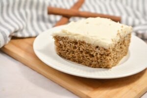 A slice of moist Keto Spice Cake topped with creamy frosting sits on a white plate, placed on a wooden cutting board. In the background, a striped cloth and two whole cinnamon sticks add a rustic touch to the scene.