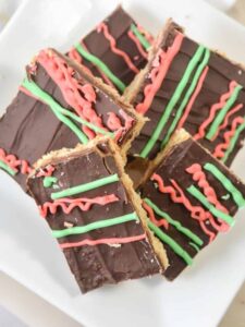 A stack of Keto Christmas Crack topped with green and pink frosting drizzles on a white plate. The bars have a smooth chocolate layer with diagonal lines of brightly colored icing.