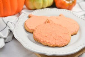 A white plate holds three keto pumpkin-shaped cookies topped with keto orange frosting. In the background, there are various decorative gourds and pumpkins in orange, green, and yellow shades, contributing to a festive autumn theme.