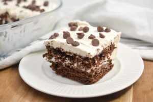 A slice of keto chocolate icebox cake sits on a white plate. The cake has layers of chocolate wafers and whipped cream, topped with a generous amount of chocolate chips. A glass dish with more of the cake is in the background, resting on a white cloth.