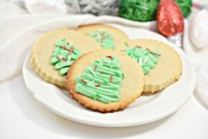 Four round keto cut-out sugar cookies with slightly browned edges are decorated with green Christmas tree-shaped keto frosting and sprinkled with red, white, and green sprinkles. The cookies are arranged on a white plate, with a hint of a green basket in the background.