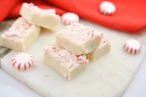 Three pieces of Keto Peppermint Fudge are stacked on a white marble surface. The fudge is topped with crushed peppermint candies, adding a festive touch. A red cloth and additional pieces of fudge are visible in the background.