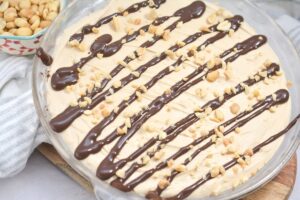 A creamy keto peanut butter pie in a dish, topped with drizzled chocolate sauce and sprinkled with chopped peanuts. A bowl of additional peanuts is partially visible to the side, along with a striped cloth napkin.