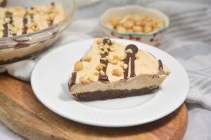 A slice of Keto Peanut Butter Pie featuring a creamy top, drizzled with chocolate and sprinkled with crushed nuts, is served on a white plate. In the background, a bowl filled with whole nuts is visible, along with the rest of the dessert on a serving dish.
