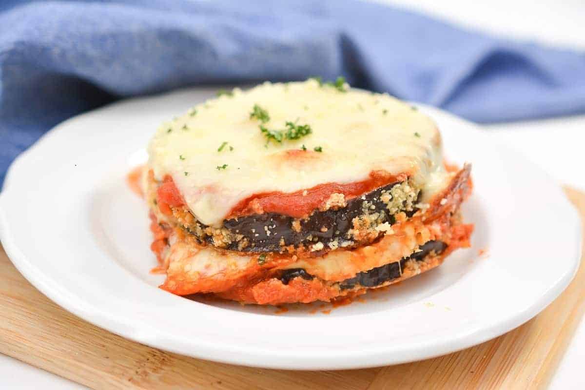 A neatly plated dish of keto eggplant Parmesan sits on a white plate. The dish features layers of breaded and fried eggplant slices, marinara sauce, and melted cheese on top, garnished with chopped herbs. A blue cloth is visible in the background.