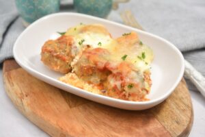 A white bowl of cheesy Keto Meatball Casserole with golden, melted cheese topping, garnished with fresh herbs, sitting on top of a wooden cutting board, ready to be enjoyed.
