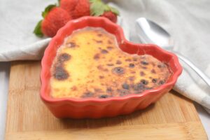 A heart-shaped ketp crème brûlée with a caramelized sugar top sits in a red ramekin on a wooden board. A silver spoon and fresh strawberries are in the background, suggesting a dessert setting.