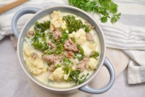 A bowl of creamy Keto Zuppa Toscana with pieces of sausage, cauliflower, and kale. A spoon is resting in the bowl, and fresh parsley is scattered in the background. The bowl sits on a white surface.