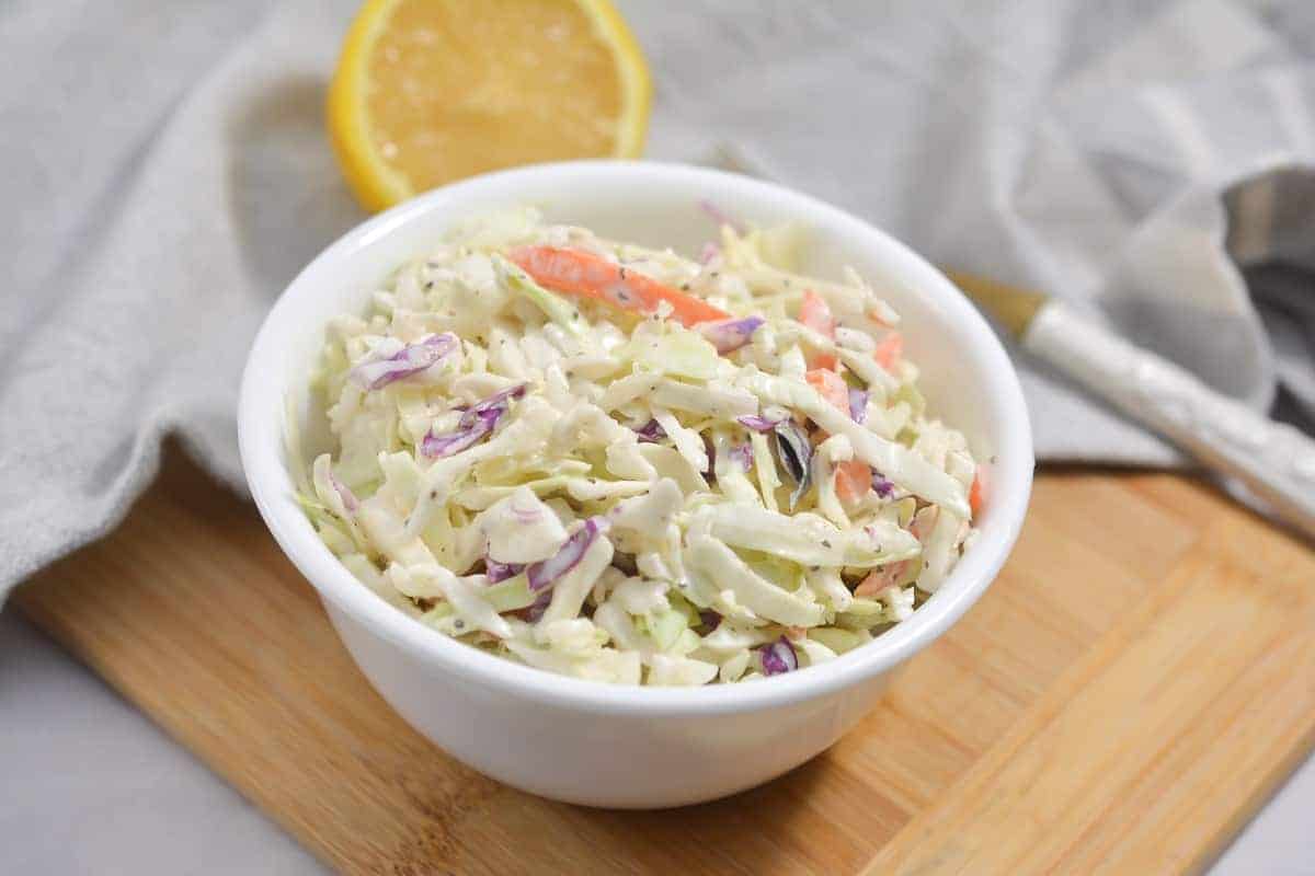 A white bowl filled with creamy keto coleslaw is placed on a wooden board. The coleslaw includes shredded green and purple cabbage, and carrot strips. A half lemon is visible in the background.