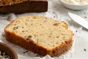 A close-up of a slice of keto seed bread with sunflower and flax seeds, resting on parchment paper. A bowl of cream cheese spread and a butter knife are in the background, adding to the rustic kitchen setting.