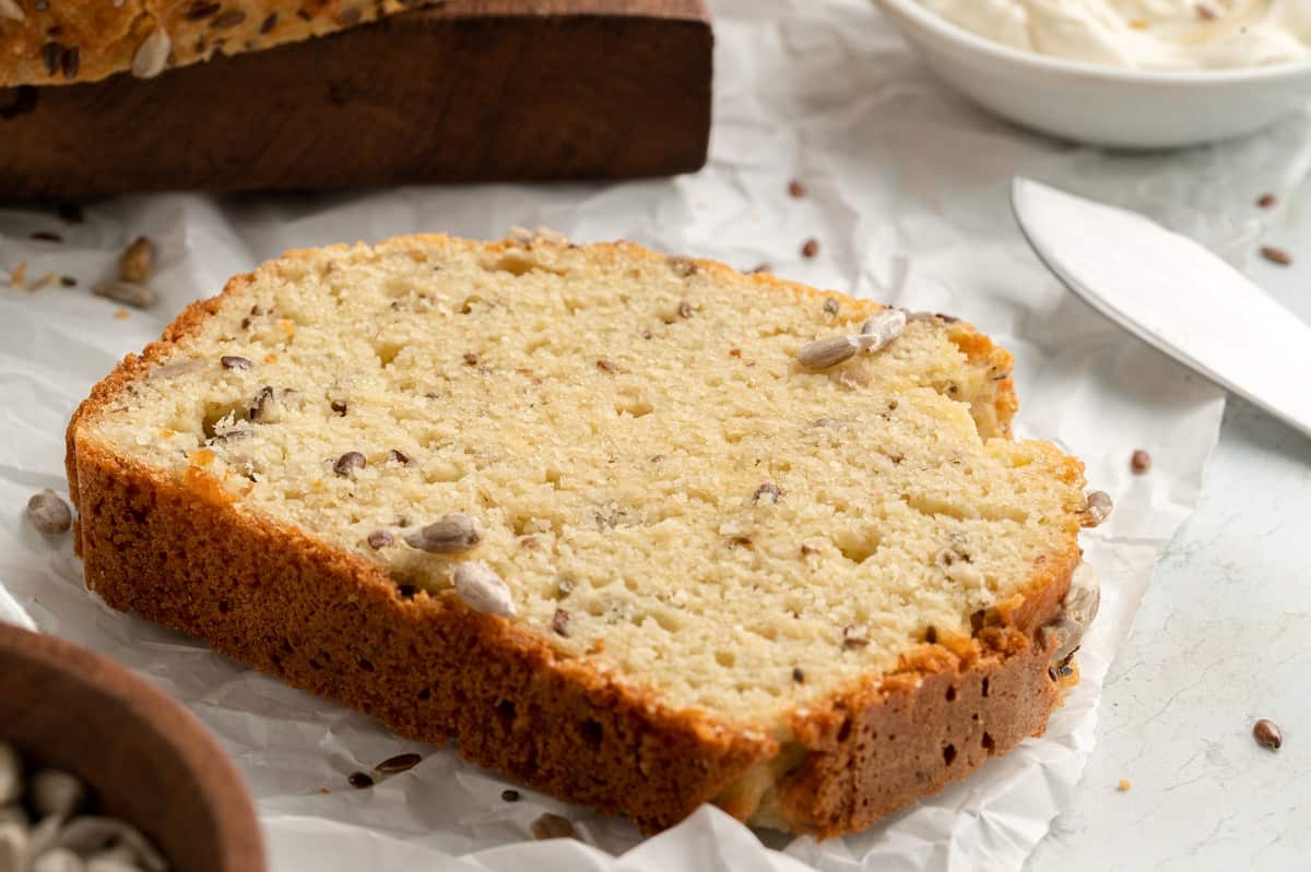 A close-up of a slice of keto seed bread with sunflower and flax seeds, resting on parchment paper. A bowl of cream cheese spread and a butter knife are in the background, adding to the rustic kitchen setting.