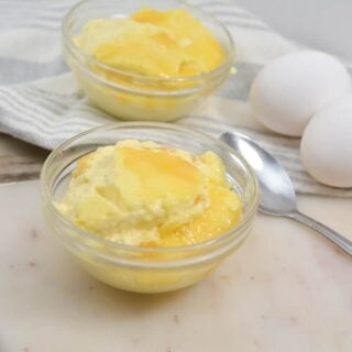 Two bowls of creamy Keto Vanilla Custard on a wooden cutting board, with a striped kitchen towel, three whole eggs and a spoon on a marble surface.