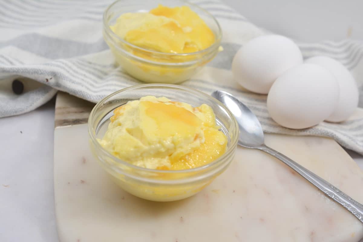 Two bowls of creamy Keto Vanilla Custard on a wooden cutting board, with a striped kitchen towel, three whole eggs and a spoon on a marble surface.