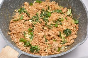 A frying pan filled with stir-fried keto dan dan noodles, ground meat, chopped green onions, and leafy greens. The dish appears to be seasoned and garnished with crushed peanuts, all mixed together. The handle of the pan is visible on the left side.