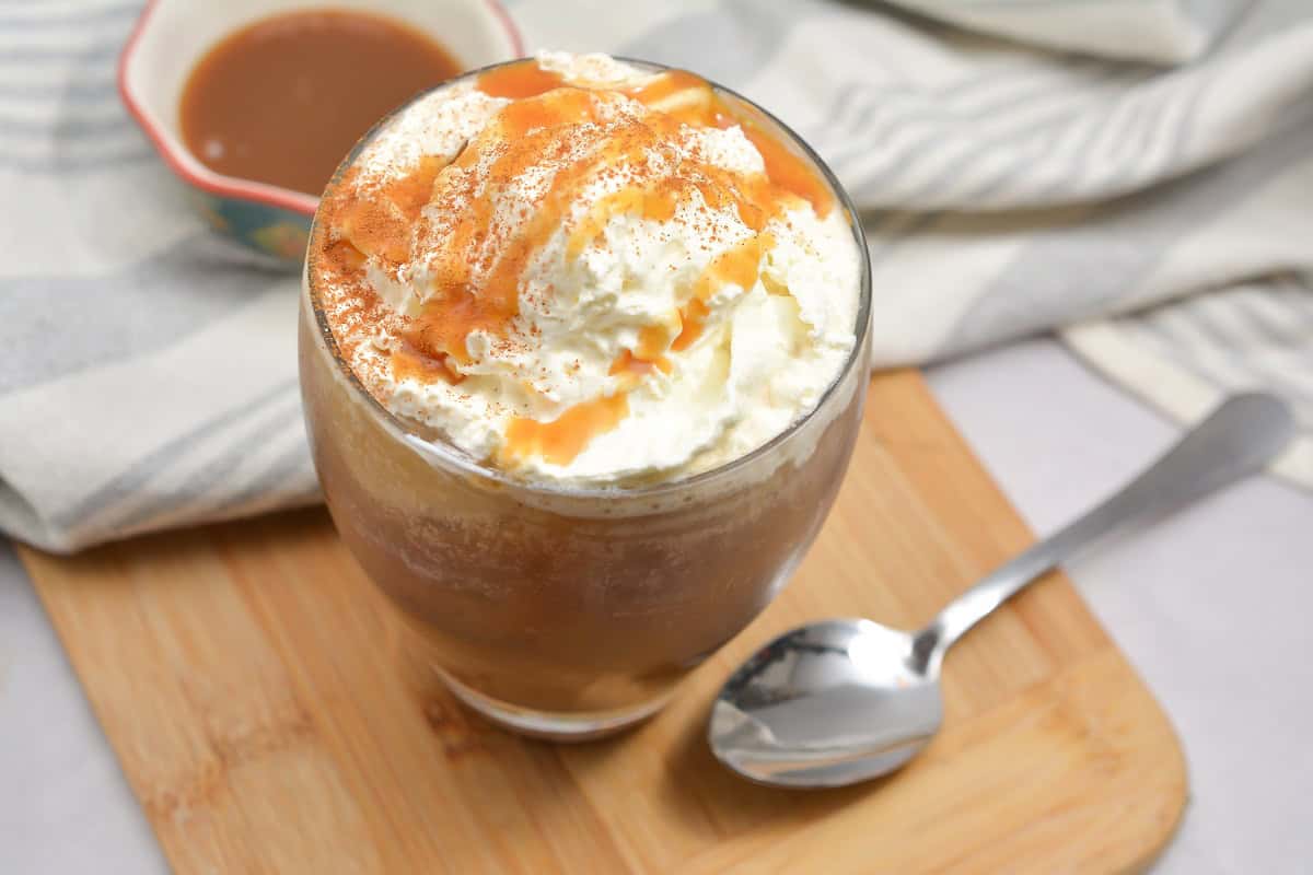 An iced Snickerdoodle Low Carb Macchiato topped with whipped cream and caramel drizzle sits on a wooden board. A spoon is placed beside the glass. In the background, there is a cup with brown liquid and a striped cloth.
