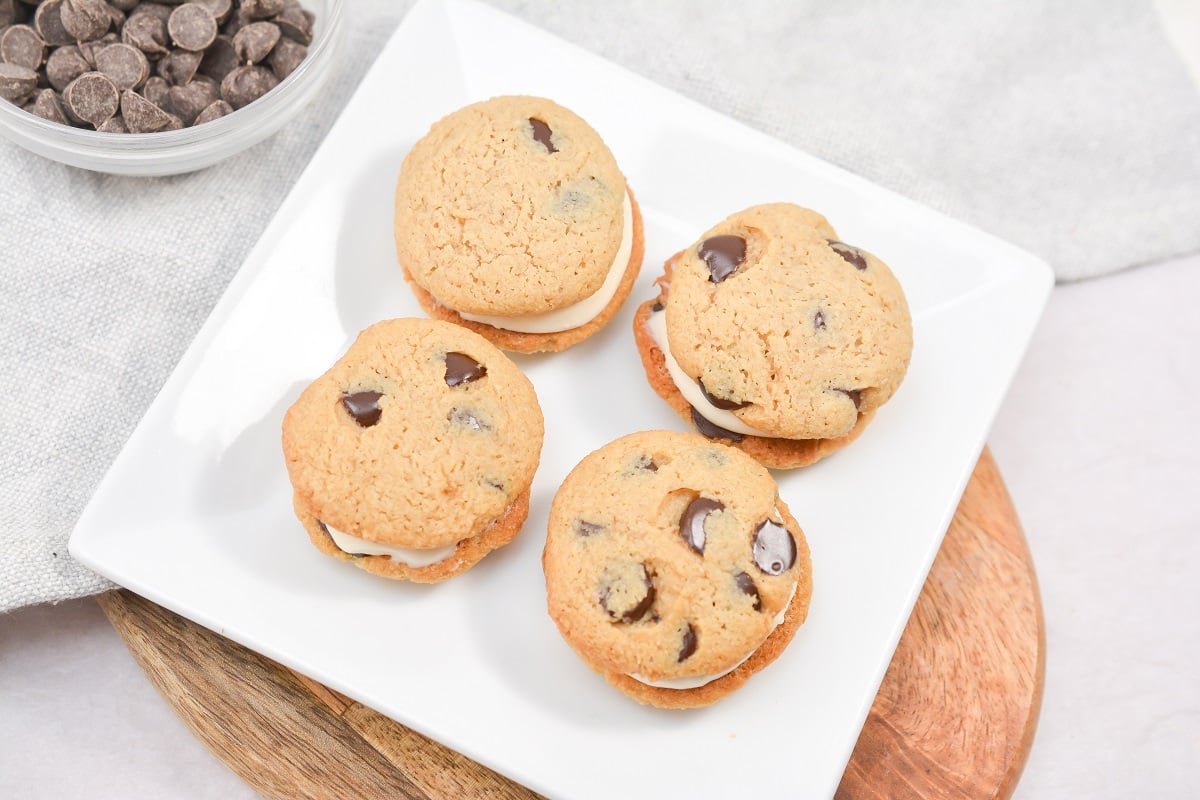 A square white plate holds four keto chocolate chip cookie sandwiches, each filled with keto icing. The plate is set on a wooden board. A small bowl of chocolate chips is placed in the top left corner of the image.