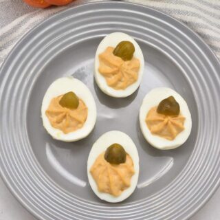A gray plate with four deviled eggs, each topped with a small gherkins pickle slice. In the background, there are two decorative gourds and a striped cloth napkin.