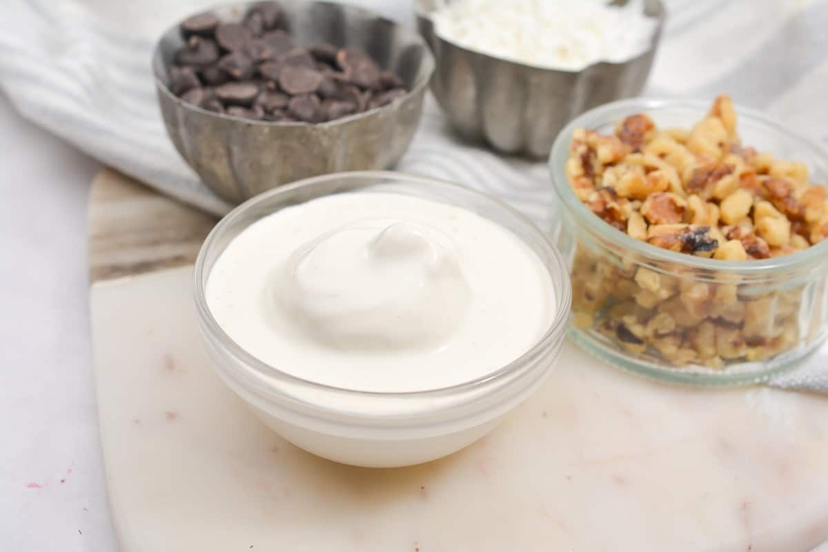 A glass bowl filled with creamy Keto Vanilla Shake is placed on a white surface. Behind it, a small glass bowl containing chopped walnuts and a metal bowl with sugar-free chocolate chips are visible. Another metal bowl, partially in view, holds shredded coconut. A striped cloth is in the background.