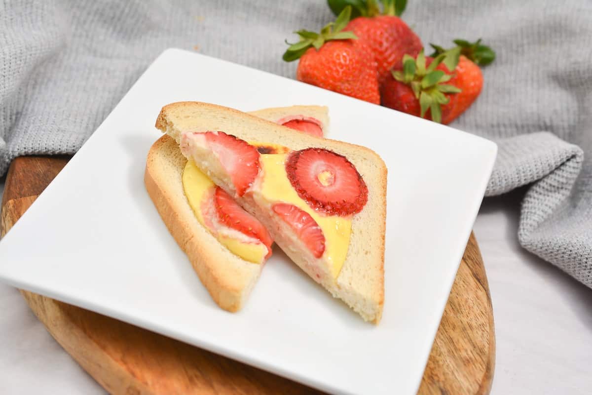 A white square plate holds keto Custard Toast, topped with a creamy custard and slices of fresh strawberries, with more strawberries to the top of the frame.