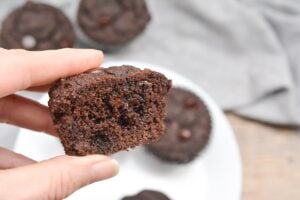 A hand holding a Chocolate Chip Keto Chocolate Muffins sliced in half, showing the moist, dense interior. Three other muffins are in the background on a white plate, with a gray cloth partially visible behind them on a wooden surface.