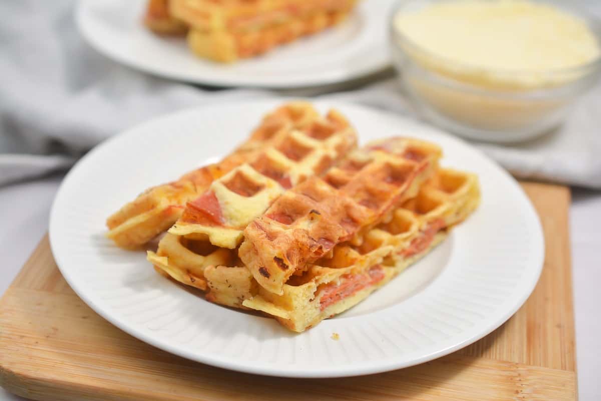 Plate of savory Keto Pizza Chaffles with chunks of peperoni and cheese, served with a bowl of Parmesan cheese in the background, on a cloth-covered table.
