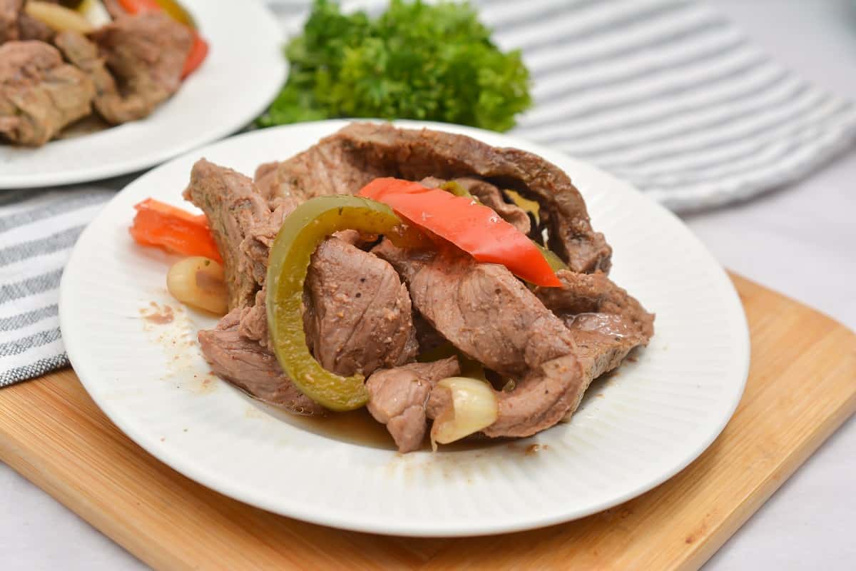 A plate of Keto Steak Fajita meat with green and red bell peppers and garlic cloves, served on a white plate with a striped cloth and parsley in the background.