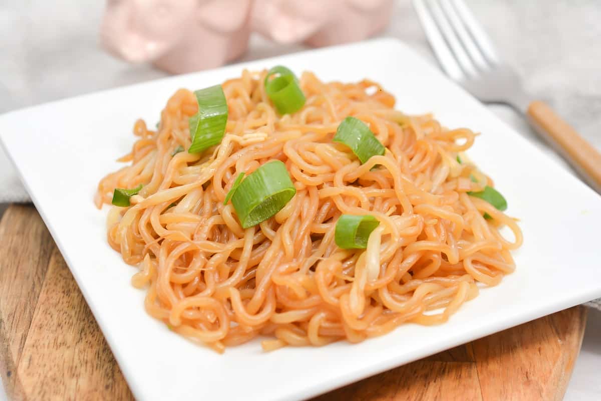A plate of keto Soy Sauce Noodles with chopped green onions served on a wooden board, accompanied by a pink pig-shaped salt and pepper shaker set and a fork on a gray napkin.