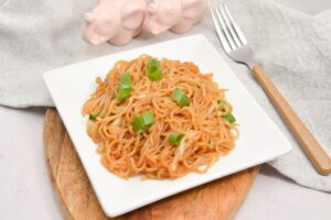 A plate of keto Soy Sauce Noodles with chopped green onions served on a wooden board, accompanied by a pink pig-shaped salt and pepper shaker set and a fork on a gray napkin.