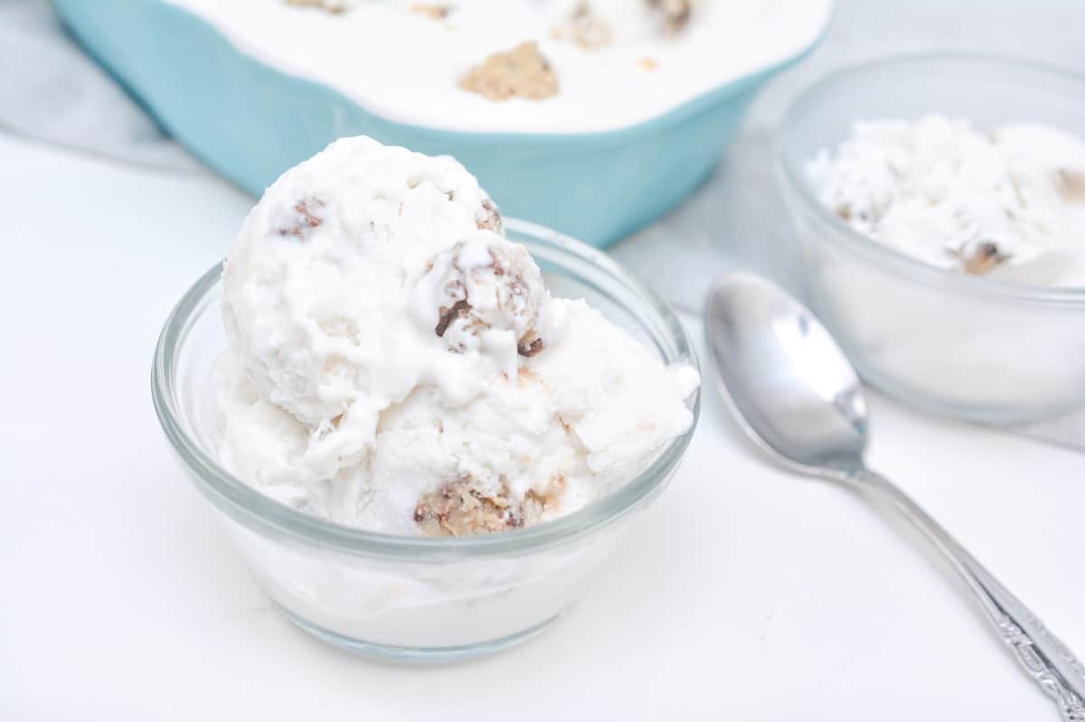 A clear glass bowl filled with Keto Cookie Dough Ice Cream containing cookie dough chunks is in the foreground. Behind it is another bowl of the same ice cream and a silver spoon. A larger dish of the ice cream is in the background.