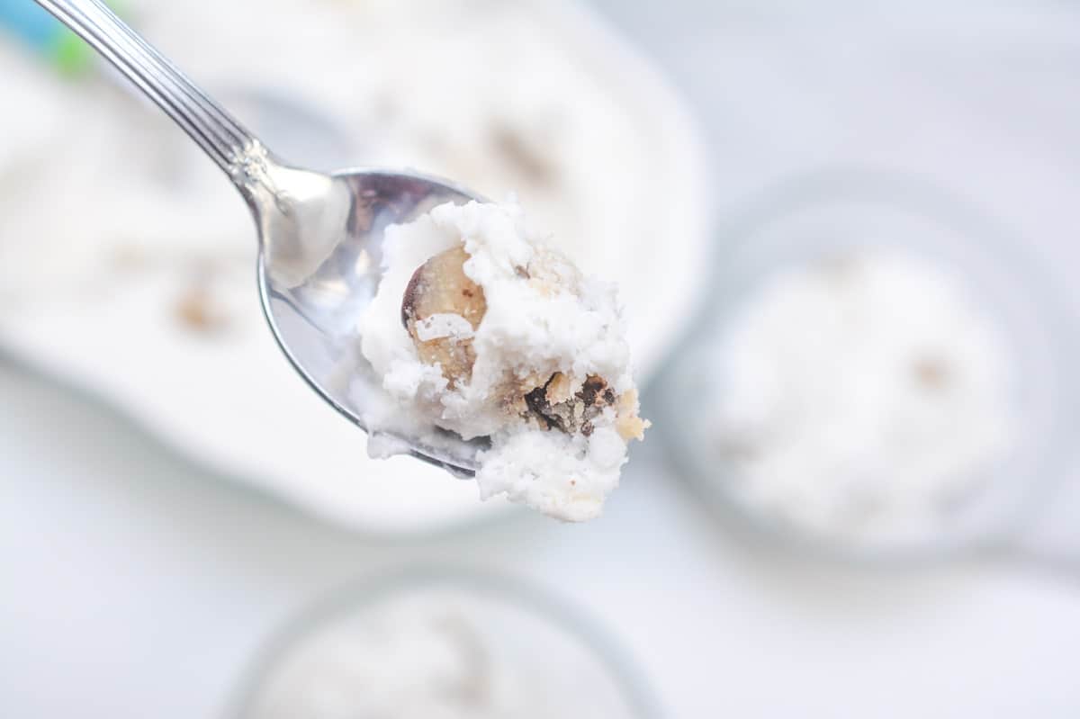 A spoon holding a bite of Keto Cookie Dough Ice Cream with visible chunks of cookie dough, against a blurry background of a bowl and two small dishes filled with more ice cream.