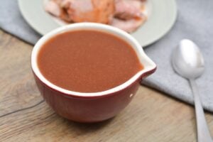 A bowl of rich, Keto Arby's Sauce on a wooden table, with a spoon to the side and a plate of sliced roast beef in the background.