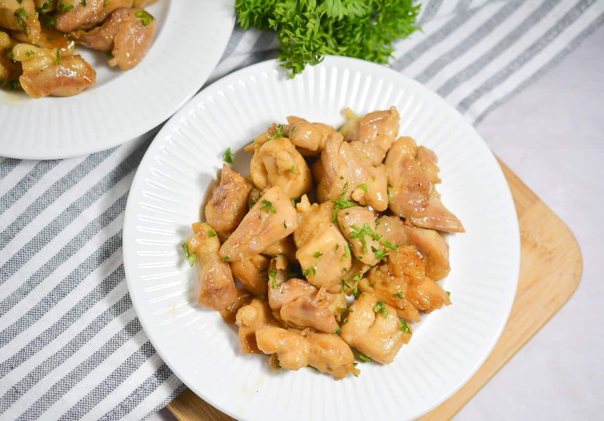 Overhead of a white plate filled with Keto Butter Chicken pieces garnished with chopped herbs is placed on a wooden cutting board. In the background, there is another plate with more chicken and a bunch of fresh parsley. The scene is set with a gray and white striped cloth.