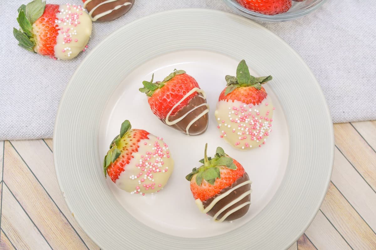 A white plate holds four strawberries dipped in white and dark chocolate, with some adorned with pink sprinkles or white chocolate drizzle. Nearby, a bowl is filled with fresh strawberries. A light gray cloth and a wooden surface are partially visible.
