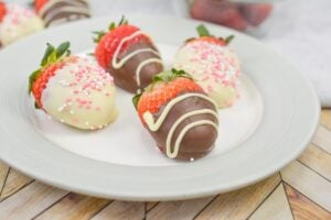 A white plate holds four strawberries dipped in white and dark chocolate, with some adorned with pink sprinkles or white chocolate drizzle. Nearby, a bowl is filled with fresh strawberries. A light gray cloth and a wooden surface are partially visible.