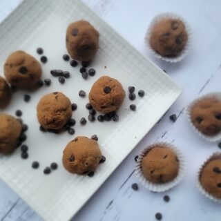 Cookies and Cream Shakeology Cookie Balls on a white square plate. Some cookies are also placed on the table with chocolate chips scattered around. A few cookies are in white paper liners. The table has a white, rustic appearance.