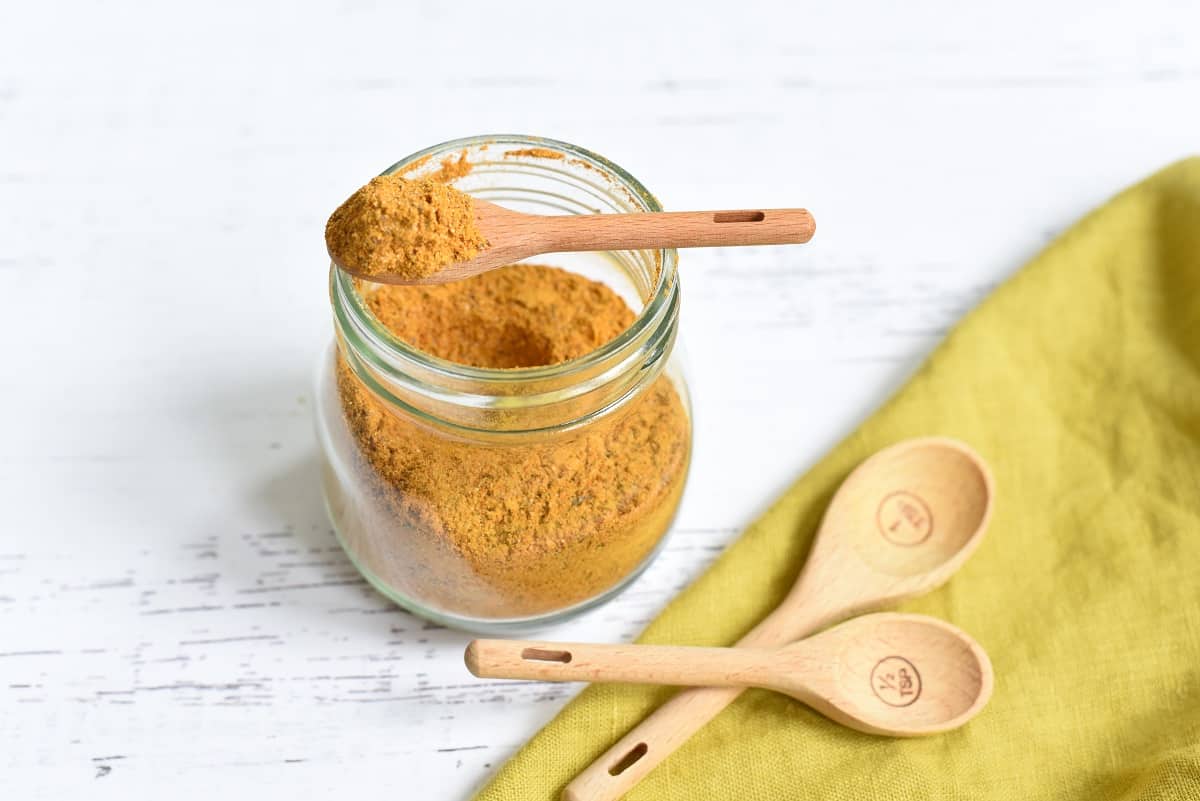 A glass jar filled with golden-brown Anti-Cancer Spice Blend powder, with a wooden spoon resting inside it. Two additional wooden spoons lie next to the jar on a white surface, with a green napkin to the side.