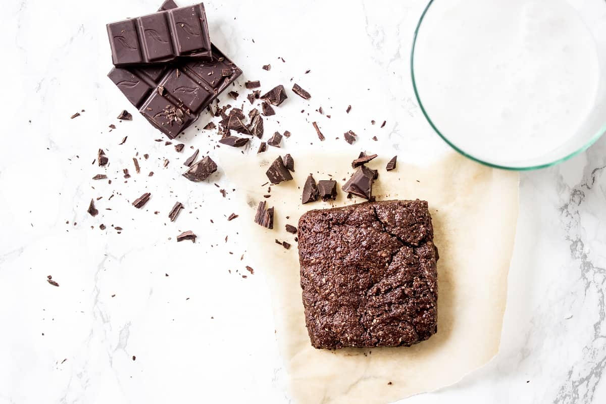 A delicious keto single-serve brownie on parchment paper, with chunks of dark chocolate beside it and a glass of milk in the corner, all set on a white marble surface.