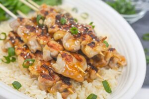 Overhead of a plate of Keto Teriyaki Chicken Skewers over cauliflower rice, garnished with green onions and sesame seeds. A small glass bowl containing more sliced green onions sits at the top of the frame.