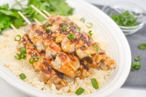 A plate of Keto Teriyaki Chicken Skewers over cauliflower rice, garnished with green onions and sesame seeds. A small glass bowl containing more sliced green onions sits at the top of the frame.