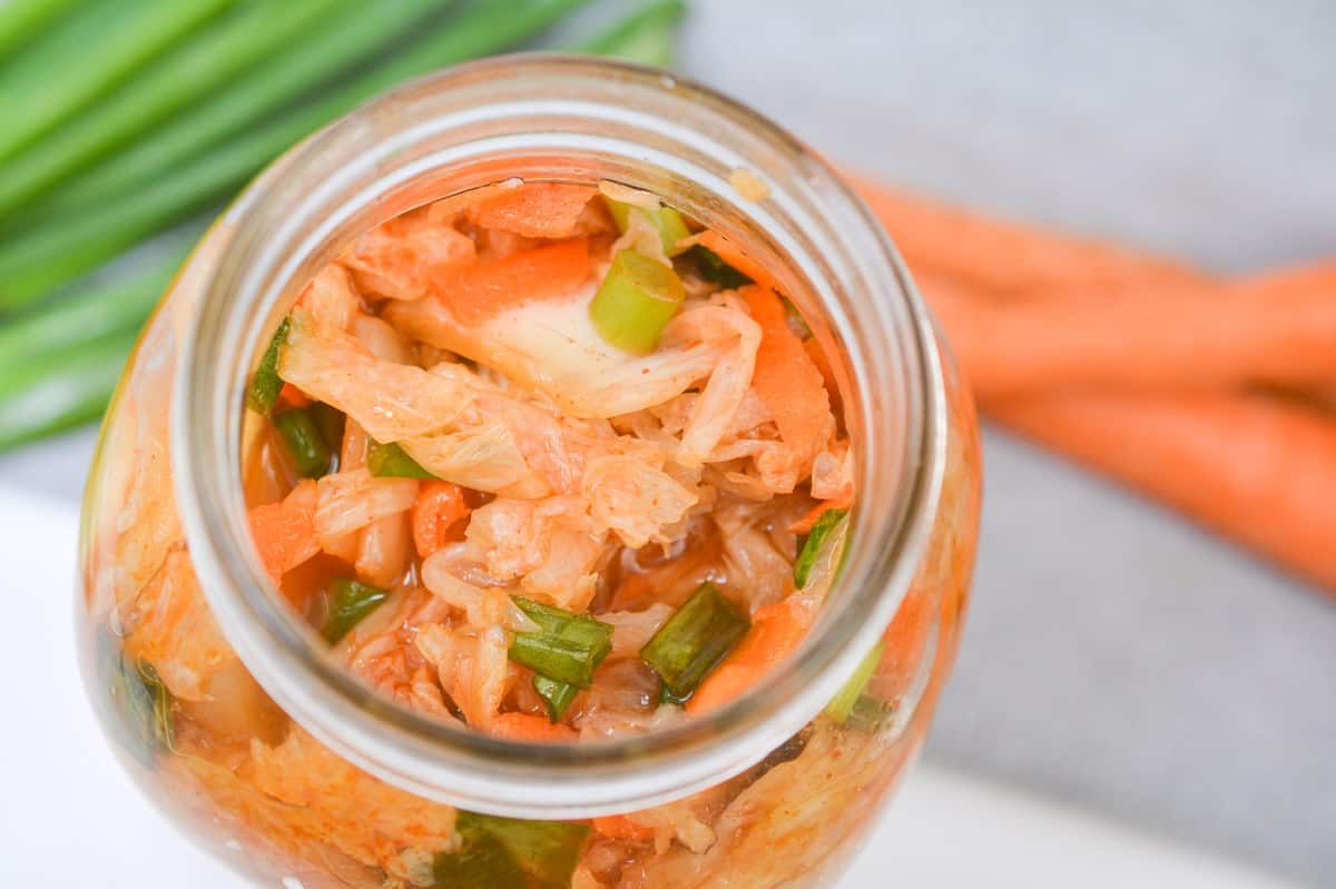 Keto Kimchi out of a glass jar, with fresh green onions and carrots in the background. The focus is on the texture and vibrant colors of the fermented cabbage.
