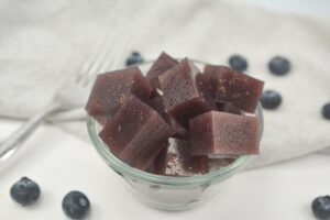 A bowl of Keto Mixed Berries Jello, placed on a white surface with scattered blueberries and a gray napkin in the background.