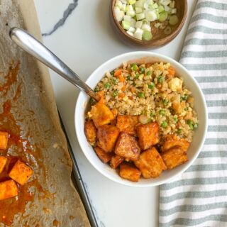 A bowl of Spicy Tofu with Fried Cauliflower Rice with diced vegetables and extra glazed tofu cubes, on a parchment lined sheet pan. Accompanied by a bowl of chopped green onions and a striped napkin on a marble surface.