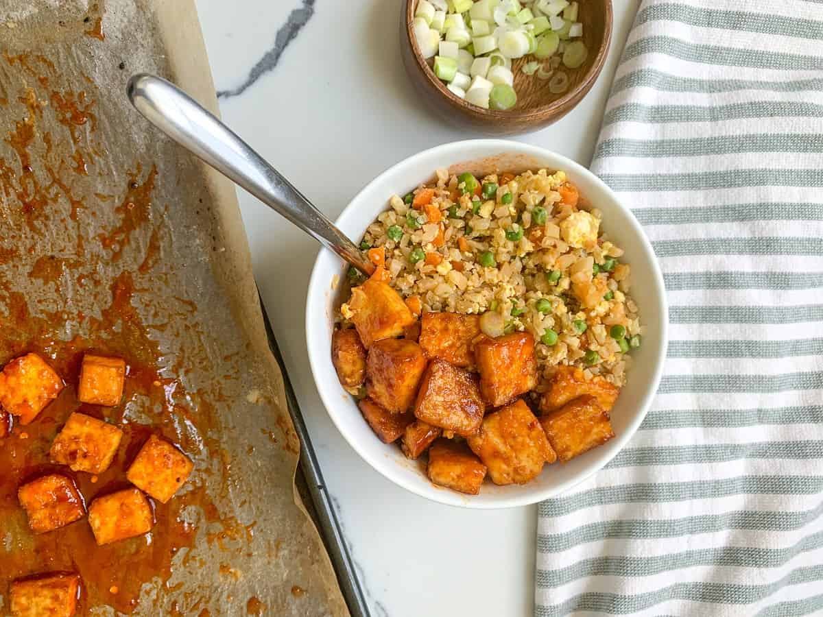 A bowl of Spicy Tofu with Fried Cauliflower Rice with diced vegetables and extra glazed tofu cubes, on a parchment lined sheet pan. Accompanied by a bowl of chopped green onions and a striped napkin on a marble surface.
