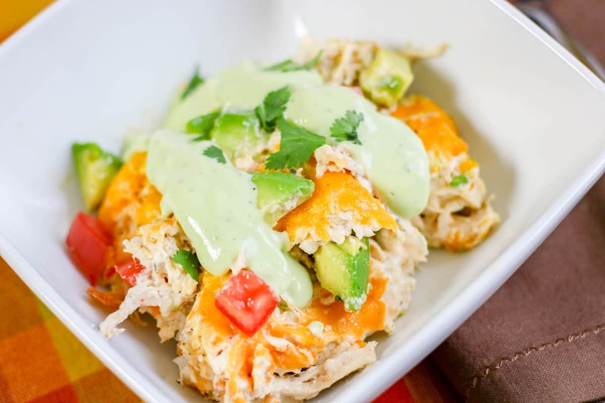 A bowl of Keto Chicken Casserole topped with melted cheese, diced tomatoes, avocado slices, and a creamy Ranch Avocado Sauce, garnished with cilantro leaves.